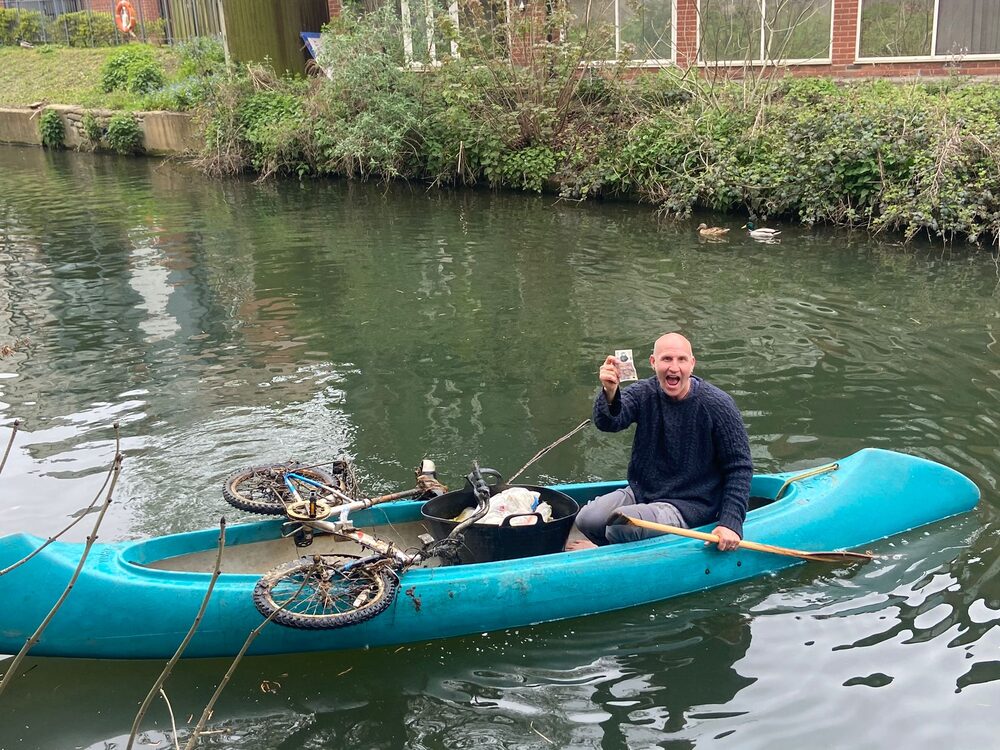 Simon Baker holding up a five pound note found during a River Stort cleanup, with a recovered bike in his canoe